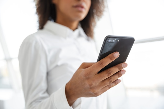 Porträt einer jungen schönen afroamerikanischen Frau mit weißem Hemd, die Handy im hellen Raum hält