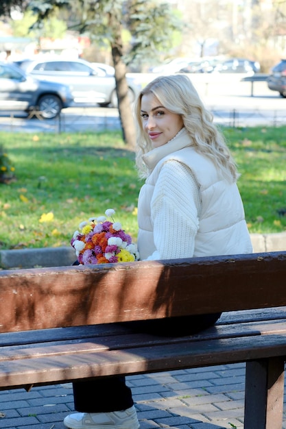 Porträt einer jungen schönen Blondine mit langen Haaren in einem weißen Pullover und Jacke im sonnigen Herbst in einem Stadtpark mit einem Blumenstrauß