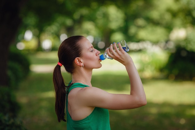 Porträt einer jungen schönen dunkelhaarigen Frau mit grünem T-Shirt Trinkwasser im grünen Park nach dem Training am Sonnenuntergang im Sommer