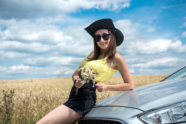 Porträt einer jungen schönen Frau, die in der Nähe ihres Autos auf einer Landstraße steht Träumen Sie von einer perfekten Reise im Sommer