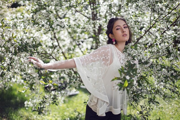 Porträt einer jungen schönen Frau in weißer Kleidung, die im Frühjahr neben einem blühenden Kirschbaum steht