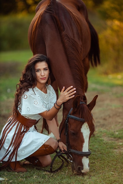 Porträt einer jungen schönen Frau mit lockigem Haar, die ein braunes Pferd hält Wandern mit den Tieren auf dem Bauernhof Reiter im Boho-Stil