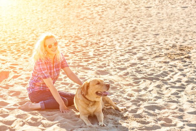 Porträt einer jungen schönen frau mit sonnenbrille, die am sandstrand mit golden retriever-hundemädchen sitzt...