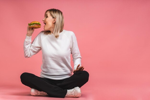 Porträt einer jungen, schönen, hungrigen Frau, die Croissant-Sandwich isst. Lokalisiertes Porträt der Frau mit Schnellimbiß über rosa Hintergrund. Diät-Konzept. Im Lotussitz auf dem Boden sitzen.