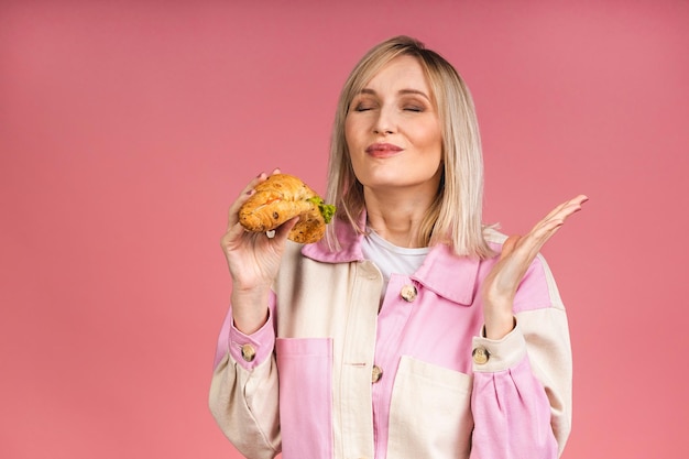 Porträt einer jungen, schönen, hungrigen Frau, die Croissant-Sandwich isst. Lokalisiertes Porträt der Frau mit Schnellimbiß über rosa Hintergrund. Diät-Konzept.