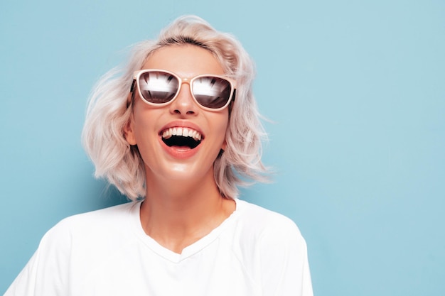 Porträt einer jungen, schönen, lächelnden Frau in trendiger, weißer Sommerkleidung. Sexy sorglose blonde Frau, die in der Nähe der blauen Wand im Studio posiert. Positives Modell, das sich drinnen amüsiert. Fröhlich und glücklich mit Sonnenbrille