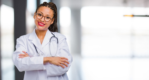 Porträt einer jungen schwarzen Doktorfrau, die seine Arme kreuzt, lächelt und glücklich und überzeugt und freundlich ist