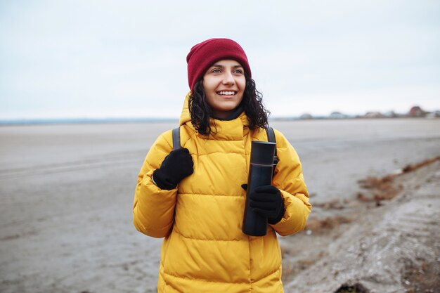 Porträt einer jungen Touristin mit einem Rucksack, der auf der Seitenstraße zwischen dem riesigen leeren Wintertal-Tiefland geht. Weiblicher Reisender, der gelbe Jacke und roten Hut trägt. Trampen, Reisekonzept.