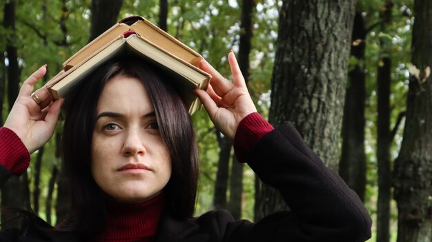 Porträt einer jungen und lustigen Brünette im Park, die ein offenes Buch auf dem Kopf hält. Lernen macht Spaß. Frau, die mit Büchern auf ihrem Kopf balanciert. Der Schüler ist müde vom Lesen.