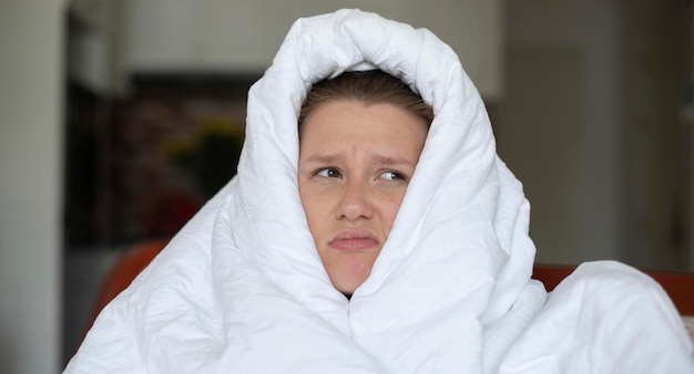 Porträt einer jungen, unglücklichen, depressiven, schönen, faulen, einsamen Frau, die sich in einer Decke bedeckt, die an Depressionen leidet, mit traurigem, verärgertem, frustriertem Blick zu Hause im Wohnzimmer am frühen Morgen