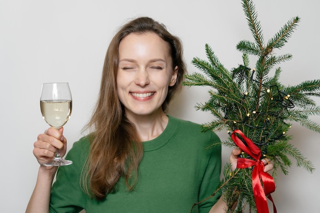 Porträt einer jungen zufriedenen Frau mit grünem Weihnachtsbaum und Wein in den Händen auf weißem Hintergrund