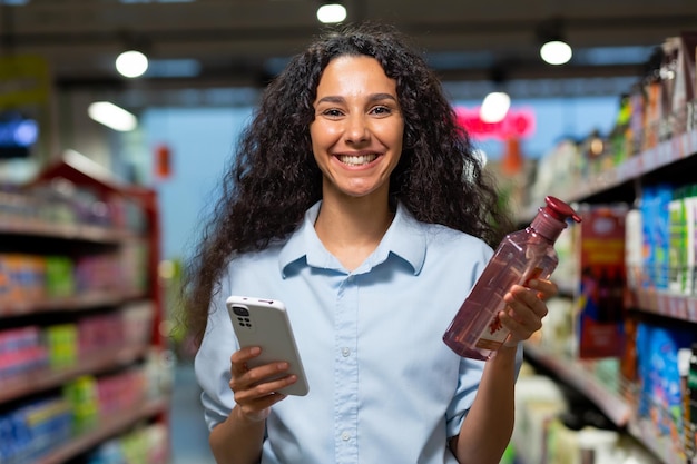 Porträt einer Käuferin in einem Supermarkt eine hispanische Frau wählt Shampoo für Haare und Duschgel, liest Rezensionen im Internet, lächelt und benutzt eine Anwendung auf ihrem Handy