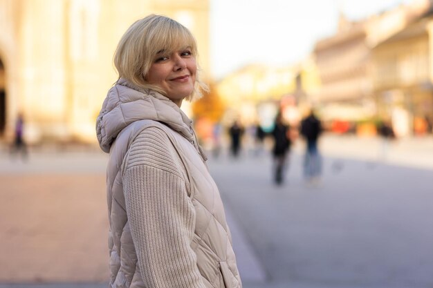 Porträt einer kaukasischen erwachsenen lächelnden blonden Frau in einer Jacke, die im Stadtzentrum posiert, Konzept der Sorglosigkeit