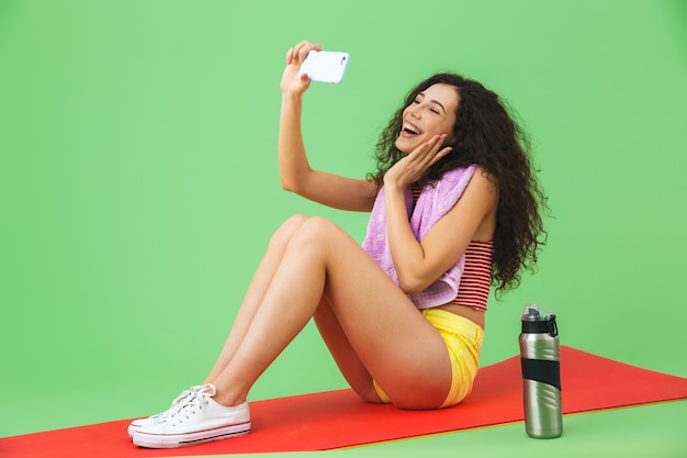 Porträt einer kaukasischen Frau der 20er Jahre in Sportkleidung mit Handtuch am Hals, die ein Selfie-Foto macht, während sie nach dem Training auf der grünen Wand auf der Fitnessmatte sitzt