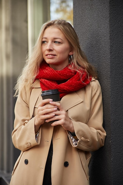 Porträt einer kaukasischen Frau im beigen Trenchcoat im Freien mit einer Tasse Kaffee