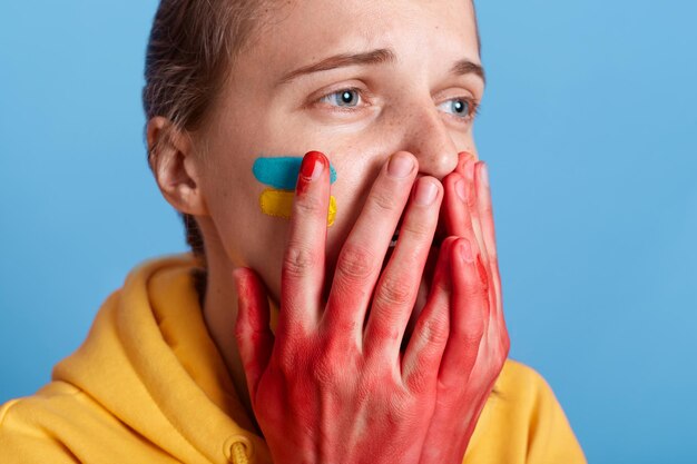 Porträt einer kaukasischen Frau mit gelbem Kapuzenpullover, die weint und den Mund mit blutigen Handflächen bedeckt, die Gewalt in der Ukraine ansieht, den Krieg stoppt und den Ruhm der Ukraine bekämpft