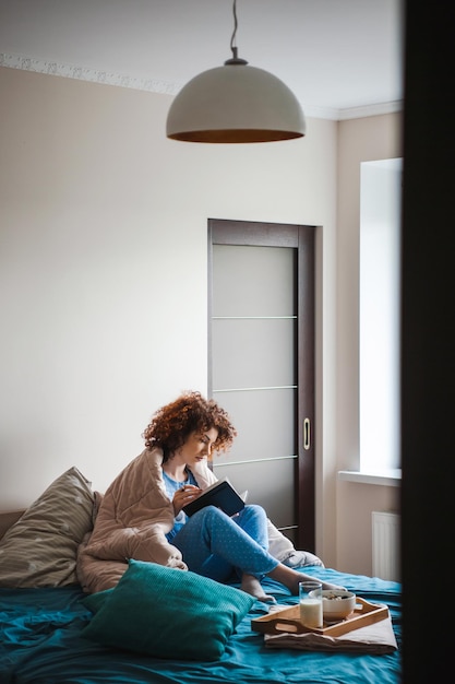 Porträt einer kaukasischen lockigen Frau, die auf dem Bett sitzt und ein gesundes Frühstück isst und intere liest