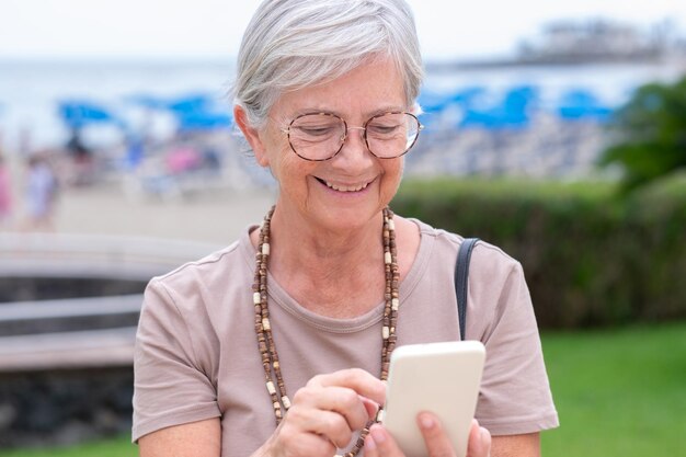 Porträt einer kaukasischen Seniorin, die an einem bewölkten Sommertag im Freien sitzt und ein Handy verwendet Attraktive lächelnde Frau, die Entspannung und Urlaub genießt