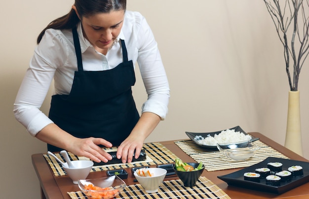 Porträt einer Köchin, die ein japanisches Sushi mit Reis, Avocado und Garnelen auf Nori-Algenblatt aufrollt