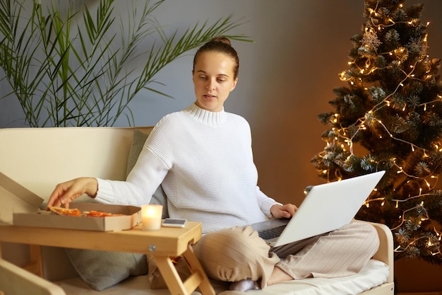 Porträt einer konzentrierten, ruhigen Frau mit braunen Haaren, die einen lässigen Pullover trägt, der zu Hause in einem dekorierten Raum für die Weihnachtsferien auf Husten sitzt und an einem Notizbuch arbeitet, das Pizza nimmt, um einen Snack zu sich zu nehmen