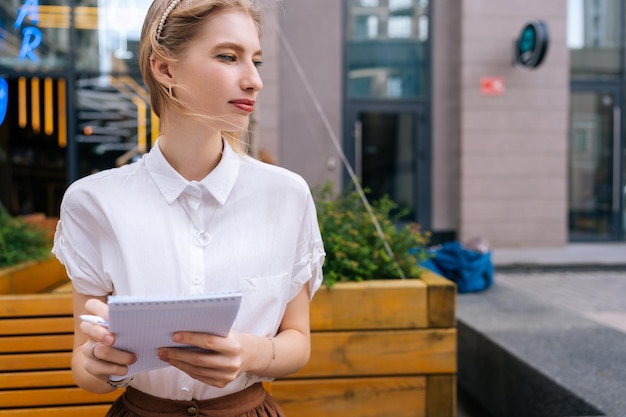 Porträt einer kreativen Künstlerin, die Zeit im Freien verbringt, indem sie ein Skizzenbuch mit leeren Seiten zum Zeichnen verwendet. Nachdenkliche blonde Frau, die sich auf der Bank neu erstellt