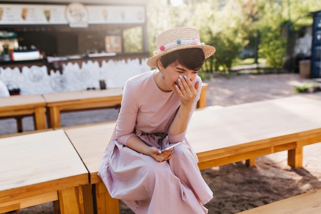 Porträt einer lachenden jungen Dame in einem langen, schönen Kleid, die auf einem Holztisch im Open-Air-Café sitzt. Anmutiges fröhliches brünettes Mädchen erhielt eine lustige Nachricht und lächelte und bedeckte das Gesicht mit der Hand.