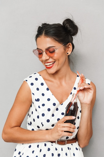 Porträt einer lachenden jungen Frau im Sommerkleid und in der Sonnenbrille lokalisiert, Flasche mit kohlensäurehaltigem Getränk haltend