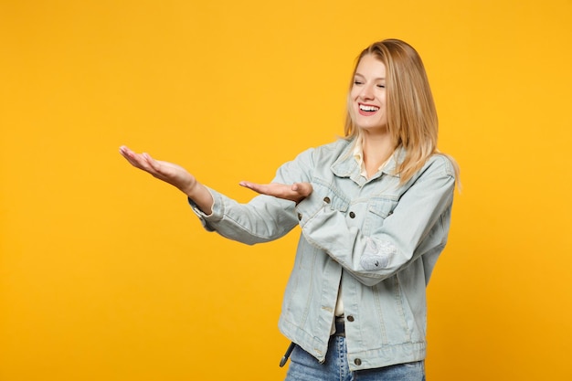 Porträt einer lachenden jungen Frau in Denim-Freizeitkleidung, die die Hände zur Seite zeigt und etwas isoliert auf gelb-orangefarbenem Wandhintergrund im Studio hält. People-Lifestyle-Konzept. Mock-up-Kopienbereich.