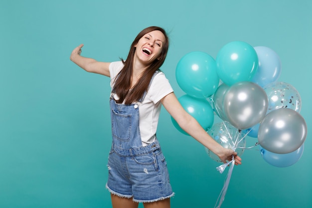 Porträt einer lachenden jungen Frau in Denim-Kleidung, die Hände ausbreitet und das Halten bunter Luftballons feiert, die auf blauem türkisfarbenem Wandhintergrund isoliert sind. Geburtstagsfeier, Menschen Emotionen Konzept.