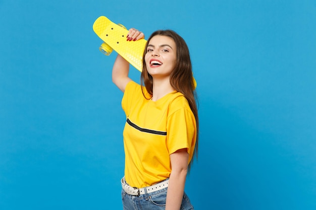 Porträt einer lachenden jungen Frau in lebhafter Freizeitkleidung, die steht, in die Kamera blickt und im Studio ein gelbes Skateboard isoliert auf blauem Wandhintergrund hält. People-Lifestyle-Konzept. Mock-up-Kopienbereich.