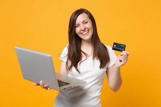 Porträt einer lachenden jungen Frau in weißer Freizeitkleidung mit Laptop-PC-Computer, Kreditkarte isoliert auf gelb-orangefarbenem Wandhintergrund im Studio. People-Lifestyle-Konzept. Mock-up-Kopienbereich.