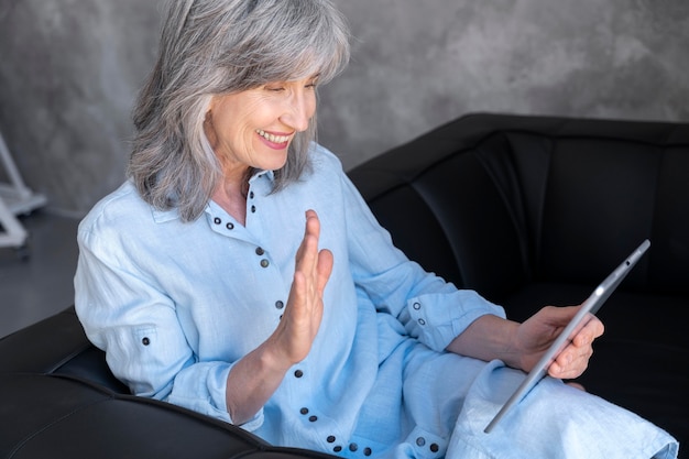 Porträt einer lächelnden älteren Frau, die zu Hause ein Tablet für einen Videoanruf verwendet und winkt