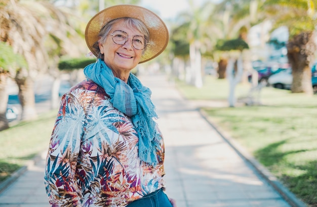 Porträt einer lächelnden älteren Frau mit Hut und Brille, die im Freien im öffentlichen Park spazieren geht und in die Kamera schaut, entspannte ältere Frau, die Freiheit und Ruhestand genießt