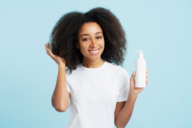 Porträt einer lächelnden afroamerikanischen Frau mit lockigem Haar in weißem T-Shirt, die eine Shampooflasche in der Hand hält