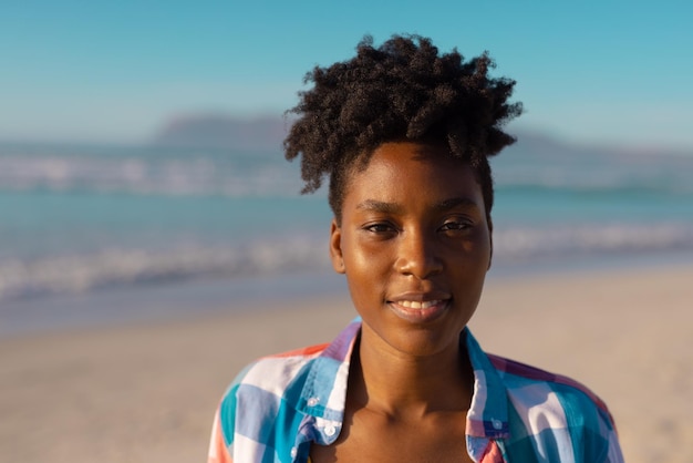 Porträt einer lächelnden afroamerikanischen jungen Frau mit kurzen lockigen Haaren am Strand vor klarem Himmel