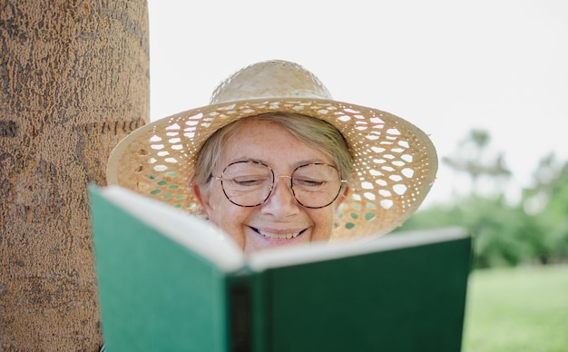 Porträt einer lächelnden alten Seniorin mit Hut und Brille, die im öffentlichen Park sitzt und ein Buch liest
