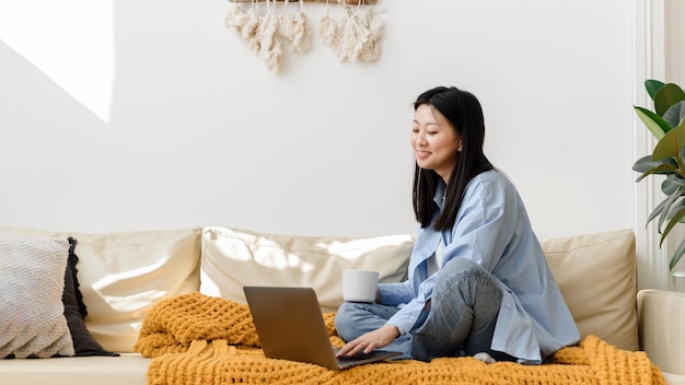 Porträt einer lächelnden asiatischen Frau, die mit einem Laptop im gemütlichen Wohnzimmer zu Hause auf dem Sofa sitzt Glückliche Studentin, die Computer für Freizeitstudien, Bildung oder Arbeit verwendet