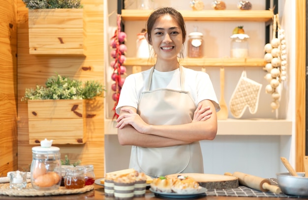 Porträt einer lächelnden asiatischen Köchin mit professioneller Schönheit, die Spaß beim Kochen mit Teig für hausgemachte Kekse und Kuchenzutaten auf dem Tisch in der Küche hat