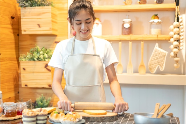 Porträt einer lächelnden asiatischen Köchin mit professioneller Schönheit, die Spaß beim Kochen mit Teig für hausgemachte Kekse und Kuchenzutaten auf dem Tisch in der Küche hat