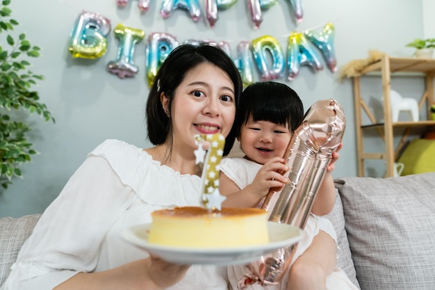 porträt einer lächelnden asiatischen mutter, die mit einem kuchen in der hand in die kamera schaut, während sie ihr glückliches kleines mädchen umarmt, das auf einer geburtstagsfeier mit einem ballonaufsteller spielt