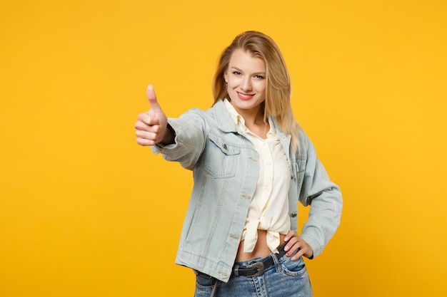 Porträt einer lächelnden, atemberaubenden jungen Frau in Denim-Freizeitkleidung, die in die Kamera schaut und den Daumen isoliert auf leuchtend gelb-orangefarbenem Hintergrund im Studio zeigt. People-Lifestyle-Konzept. Mock-up-Kopienbereich.