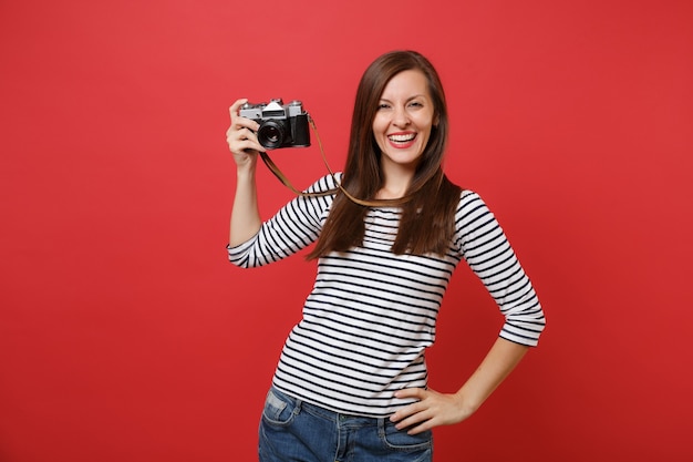 Porträt einer lächelnden, atemberaubenden jungen Frau in gestreifter Kleidung, die eine Retro-Vintage-Fotokamera isoliert auf leuchtend rotem Wandhintergrund hält. Menschen aufrichtige Emotionen, Lifestyle-Konzept. Kopieren Sie Platz.