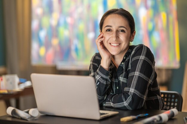 Porträt einer lächelnden Dame, die ein kariertes Hemd trägt und an einem Schreibtisch vor einem Laptop sitzt