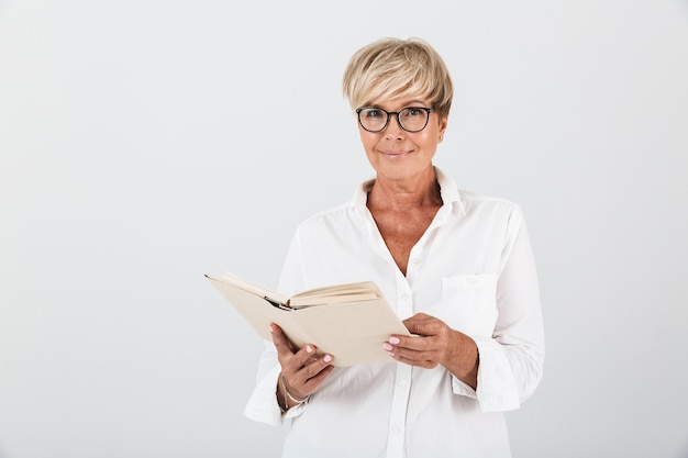 Porträt einer lächelnden erwachsenen Frau mit Brille, die ein Buch hält und die Kamera isoliert über der weißen Wand im Studio betrachtet