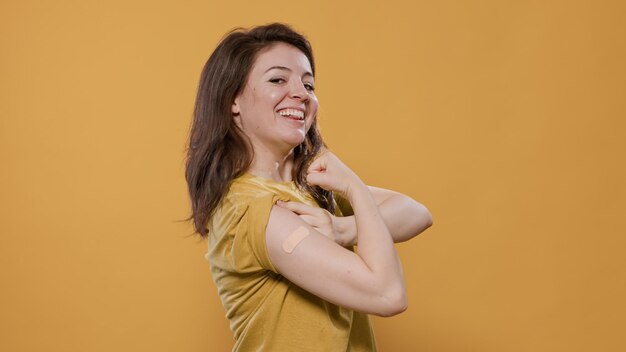 Porträt einer lächelnden Frau, die Ärmel anhebt und Pflaster nach Covid- oder Grippeimpfstoff für Gesundheitskampagnen im Studio zeigt. Stolzer junger Mensch, der einen Verband enthüllt, der die Impfstelle bedeckt.