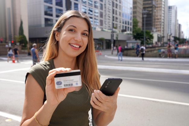 Porträt einer lächelnden Frau, die ein Smartphone hält und eine Kreditkarte zeigt, die auf die Kamera im Finanzzentrum Paulista Avenue in Sao Paulo City, Brasilien, blickt