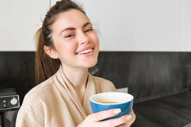 Porträt einer lächelnden Frau, die Kaffee trinkt, in der Küche steht und eine köstliche Tasse Cappuccino genießt