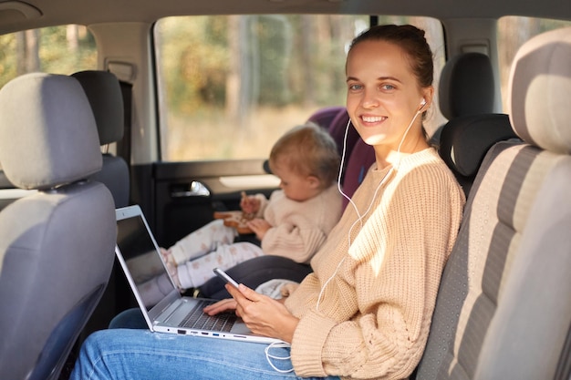 Porträt einer lächelnden Frau, die mit ihrer kleinen Tochter im Sicherheitsstuhl auf dem Rücksitz des Autos sitzt und an einem Laptop arbeitet, der ins Büro und in den Kindergarten geht und mit glücklichem Gesichtsausdruck in die Kamera schaut