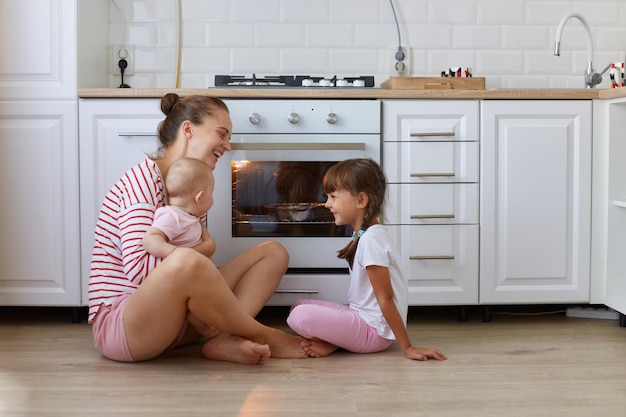 Porträt einer lächelnden Frau mit gestreiftem Freizeithemd, die auf dem Boden in der Küche sitzt, mit Kindern, die auf köstlichen Kuchen warten und mit Kindern sprechen, die Zeit miteinander verbringen