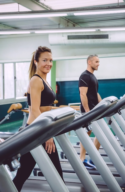 Porträt einer lächelnden Frau mit Kopfhörern, die über ein Laufband im Fitnesscenter trainiert